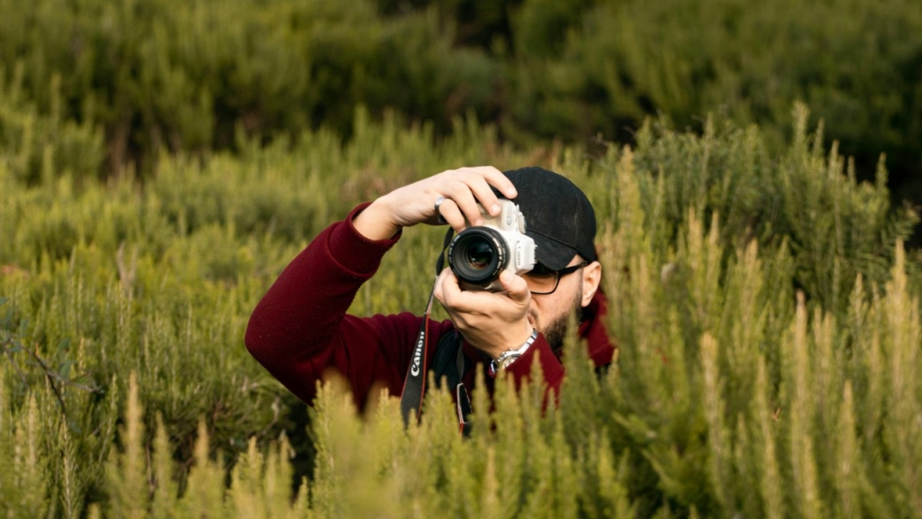 Žolėje pasislėpęs vyras fotografuoja fotoaparatu