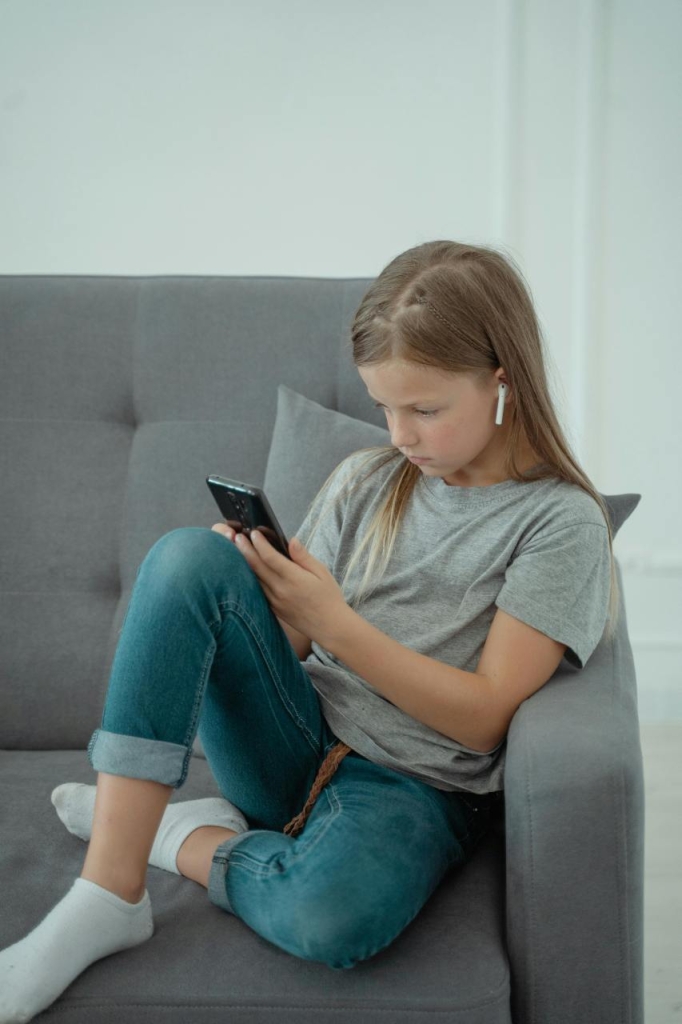 Mergaitė sėdi ant sofos ir naudojasi išmaniuoju telefonu bei belaidėmis ausinėmis