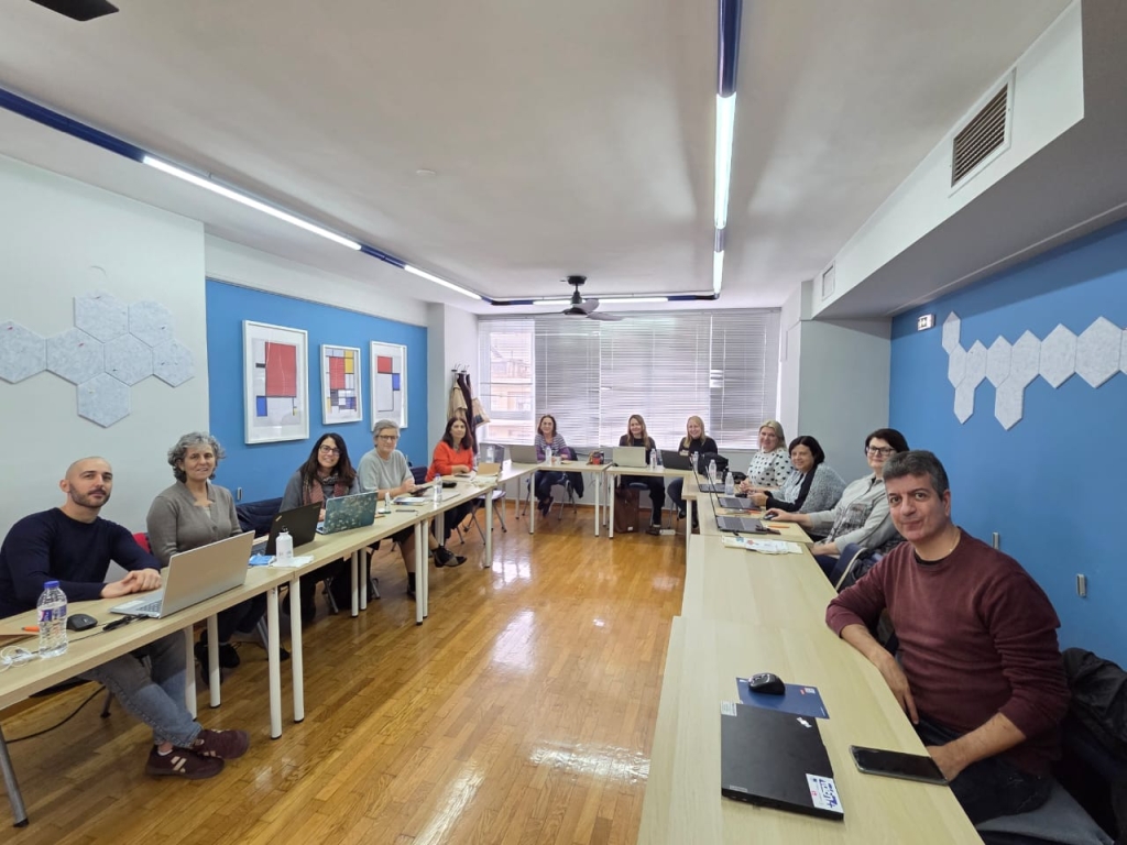 Grupė suaugusiųjų sėdi šviesioje mokymų salėje prie U formos stalų. Dalyviai turi nešiojamuosius kompiuterius.