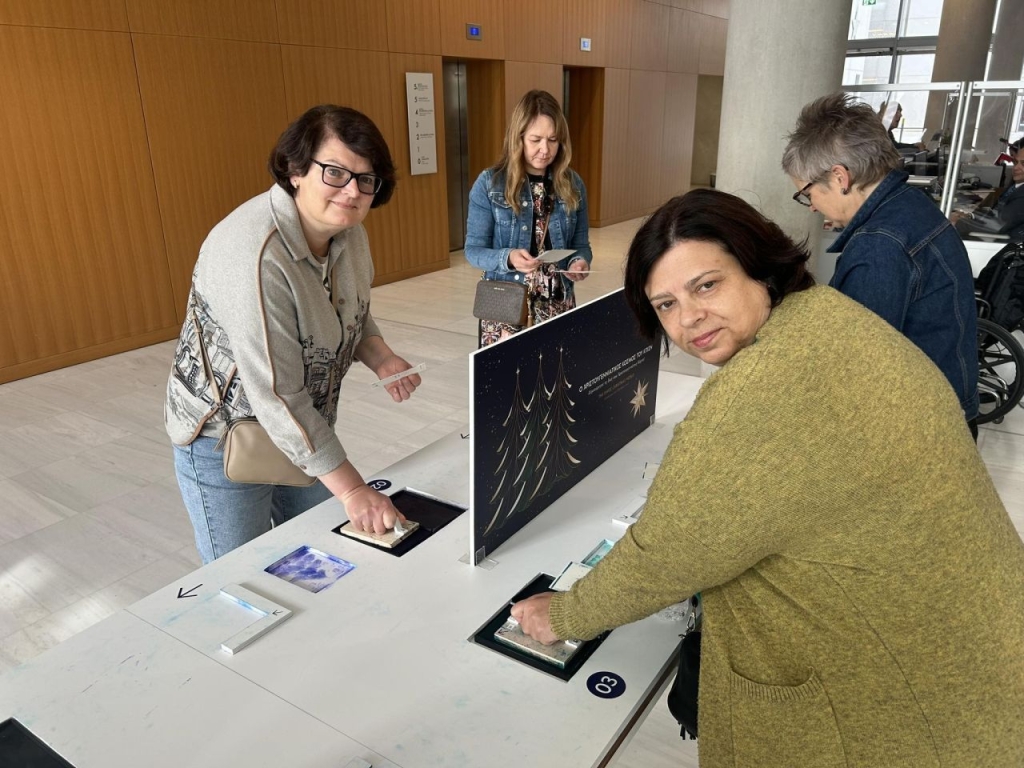 Keturi suaugę žmonės stovi moderniame viešosios erdvės interjere prie interaktyvaus stalo. Dvi moterys priekyje liečia ar spaudžia ekspozicijos elementus, įmontuotus stalviršyje, ir žiūri į kamerą. Ant stalo matyti informacinės kortelės, numeriai ir vertikalus stendas su iliustracija.