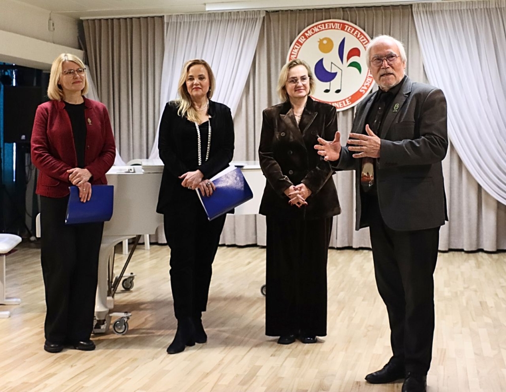 4 suaugę žmonės (3 moterys ir vyras), stovintys scenoje. Vyras kalba gestikuliuodamas rankomis.