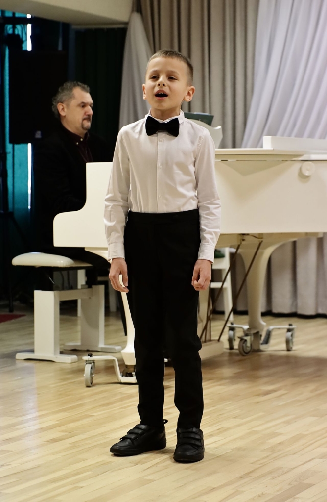 Scenoje berniukas dainuoja, už jo - pianinu grojantis vyras.