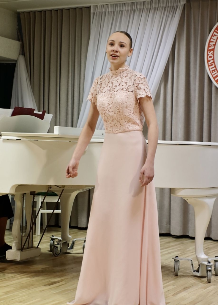 Scenoje atsistojusi dainuoja mergaitė, apsivilkusi šviesią, ilgą suknelę.