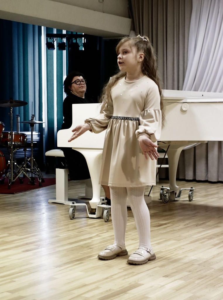 Scenoje atsistojusi dainuoja mergaitė, apsivilkusi šviesią suknelę. Už jos - pianinu grojanti moteris.