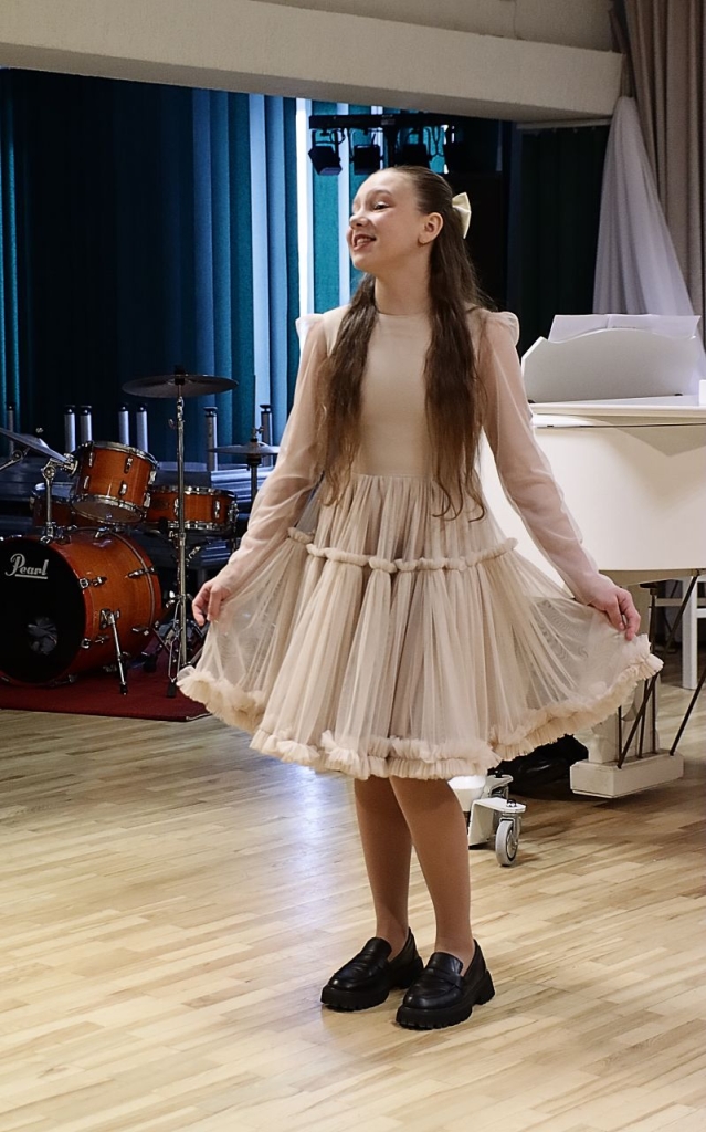 Scenoje atsistojusi dainuoja mergaitė, apsivilkusi šviesią suknelę, pritūpusi ir rankomis paėmusi suknelę.