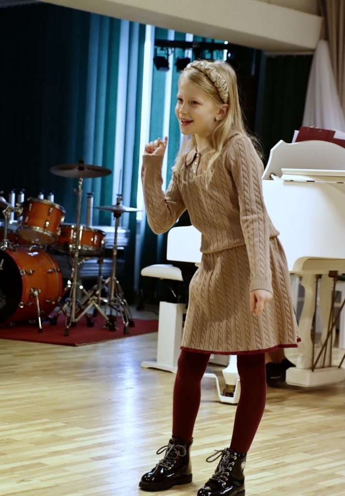 Scenoje atsistojusi dainuoja mergaitė, Ji žaismingai pasvirusi.