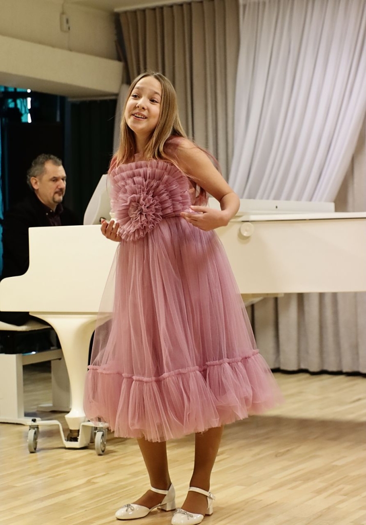 Scenoje atsistojusi dainuoja mergaitė, apsivilkusi rausvą suknelę. Už jos - pianinu grojantis vyras.