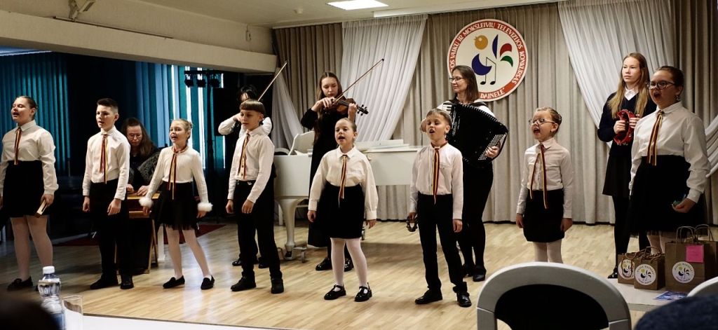 Scenoje vaikų ansamblis, kurie dainuoja. Už jų - vyresni vaikai su muzikos instrumentais.