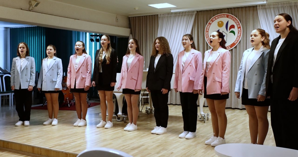 Grupė paauglių merginų scenoje, jos dainuoja.